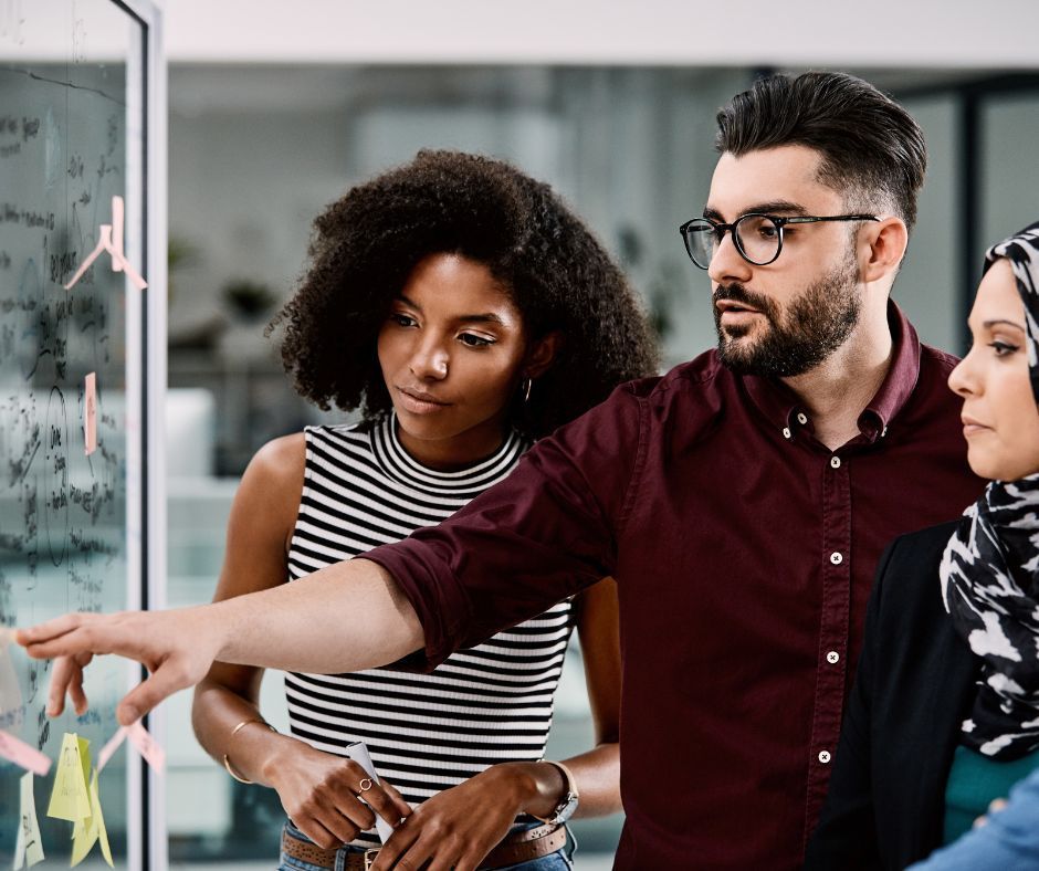 Three people looking at a glass board with notes and diagrams; man points, discussing ideas.