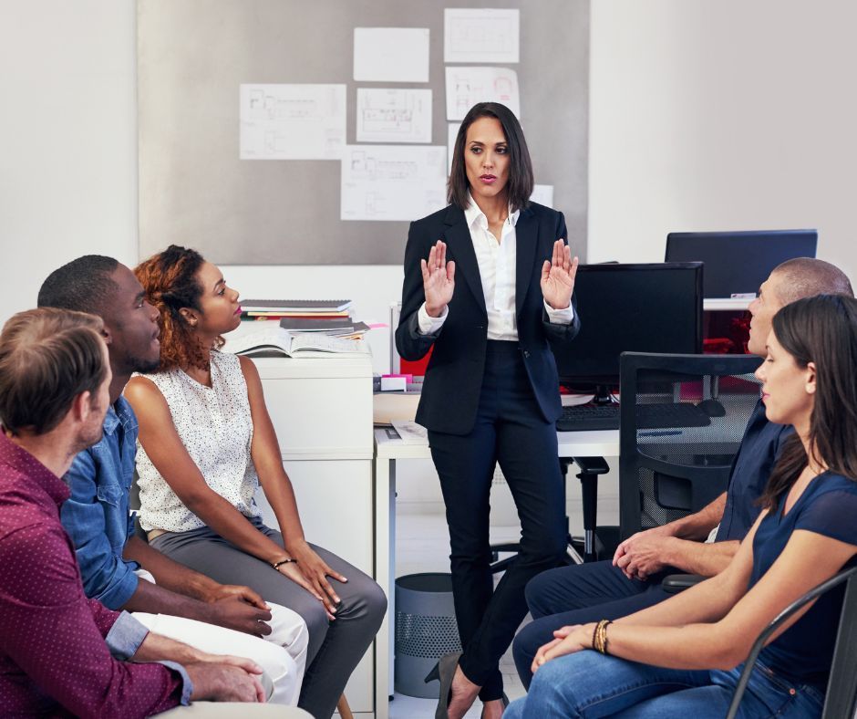 Woman in suit gesturing, addressing a seated group in an office setting.